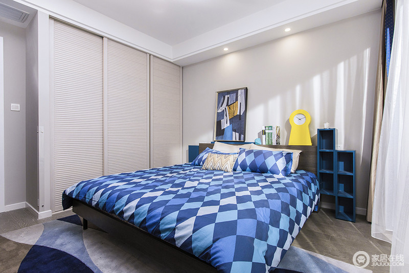 卧室以浅色漆粉刷空间，搭配定制衣柜的百叶柜门，简洁而利落；床头兼具柜子的作用，黄色时钟、蓝色抽象挂画，与菱形蓝色的床单吸睛不少，优雅之余，赋予生活能量。